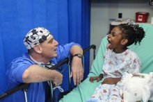 A young black girl in a hospital bed sticks out her tongue at a middle aged white man in scrubs and a surgical cap who is crouched next to the bed.