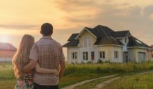 A man and a woman stand together, back to the viewer, in front of a white and yellow house.